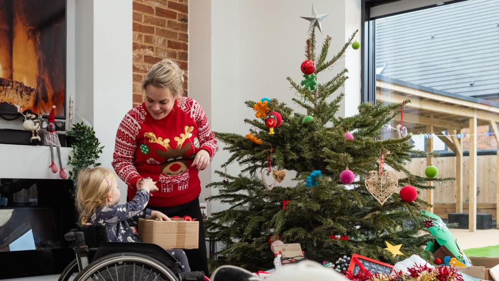 A wide angle medium close up of a young female toddler handing her mother baubles as they decorate the xmas tree together as a family. Her father and sister are sitting on the sofa. The younger daughter is sitting in her older sisters wheelchair in the living room of their family home in Newcastle upon Tyne in the North East of England