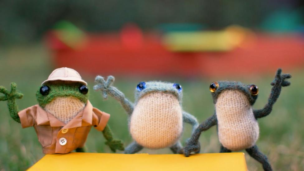 Three knitted frogs stand looking over a ledge, waving their arms.