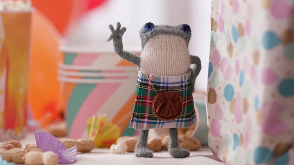 One knitted frog with blue eyes, wearing a tartan kilt around the waist, stands with one are up, in a Highland jig pose.
