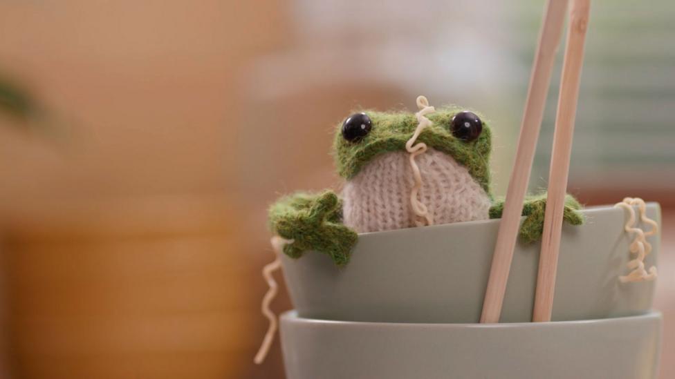 The knitted frog sits in a bowl, covered in ramen noodles.