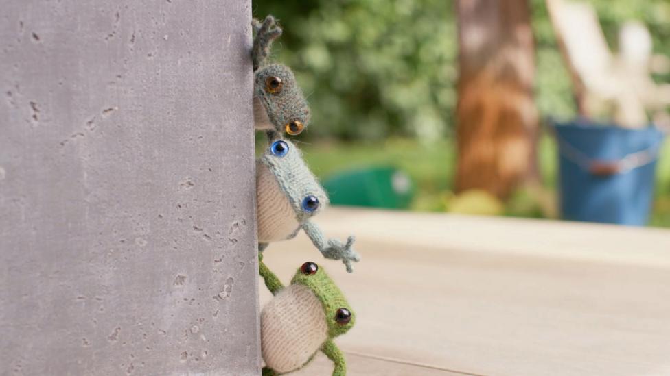 The knitted frog trio peer around a wall, stacking their heads so they can all get a look.