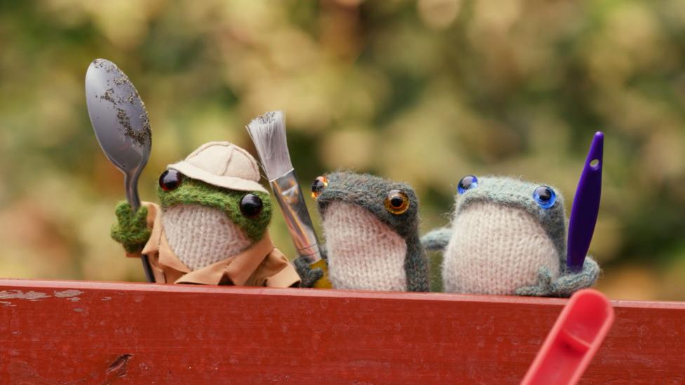 The knitted frog trio peer over a wall, holding tools such as a spoon and paintbrush.
