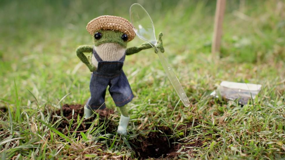 One knitted frog with dark brown button eyes wearing overalls holds a plastic spoon, and looks down on a hole dug into a lawn.