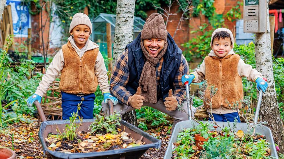 Aaron and two kids help tidy the garden with wheel barrows filled with leaves.