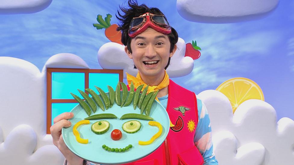 Patch, a goofy presenter wearing flight goggles and a red flight vest, holds a plate filled with vegetables in the shape of a face.