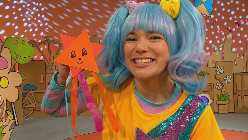 Sketch, a blue haired woman with pony tails, holds up a paper star, trailing strips.