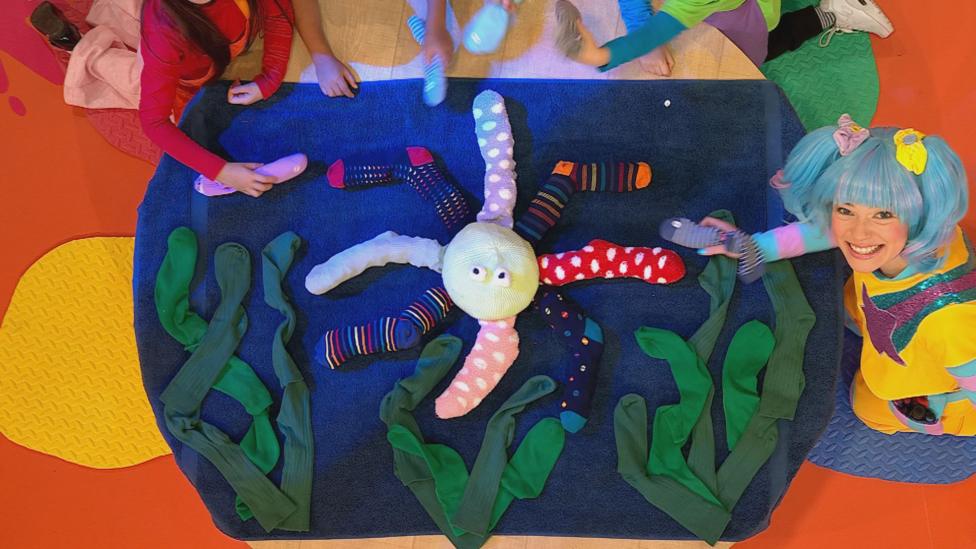 Five children gather around a birds-eye view of a table, placing 8 stuffed socks (octopus arms) onto an octopus body.