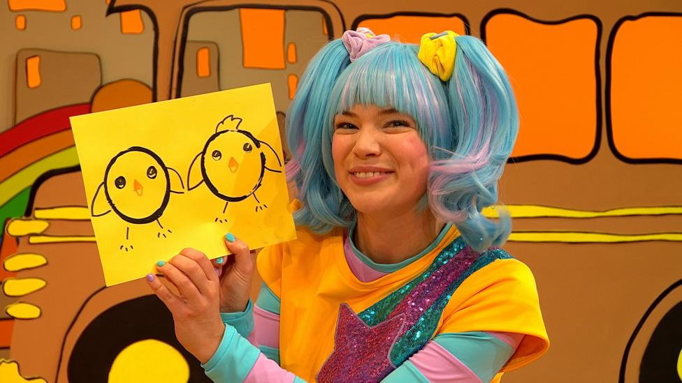 Sketch, a blue-haired woman with pigtails, smiles and holds up a piece of yellow card, containing two circular rings in paint, decorated to look like round fat birds.