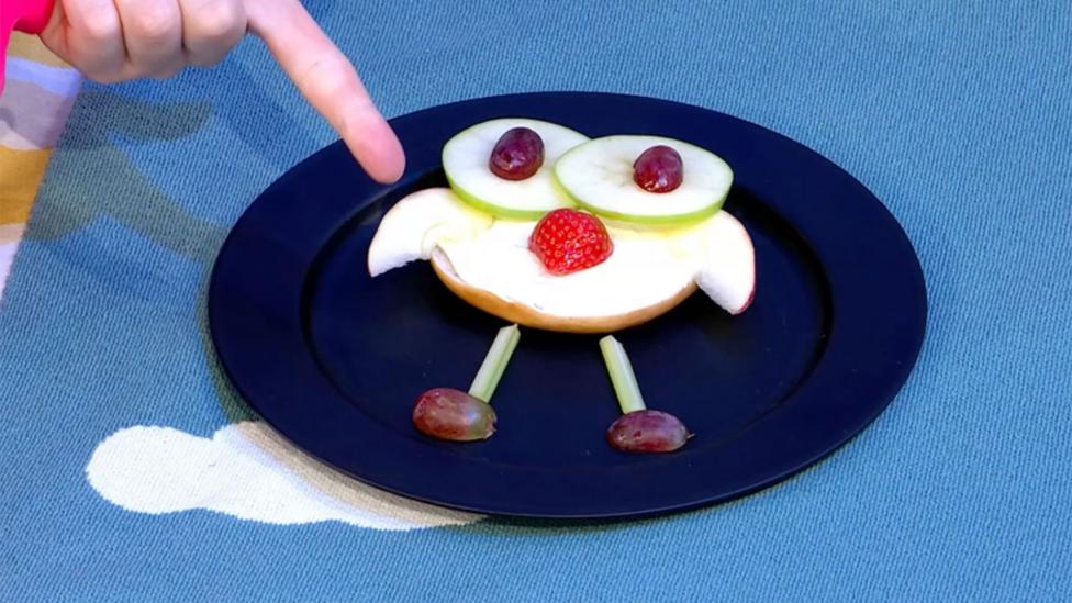 A plate holding half a bagel covered in cream cheese, with celery legs, grape feet and apples slices for eyes and wings.