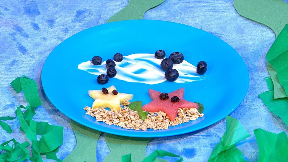 A blue plate, with star shaped cut outs of melon for starfish, blueberries as bubbles, granola sand and yoghurt waves.