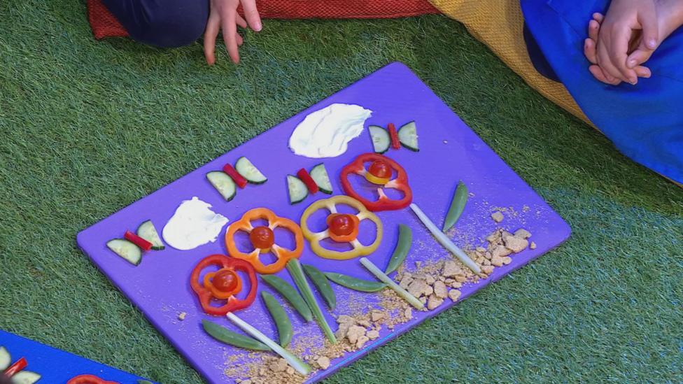 A chopping board covered in celery stalk flowers with red pepper petals, oatcake soil and cucumber butterflies.
