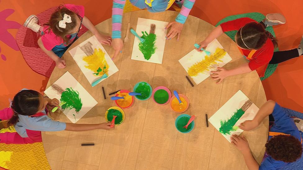Five children gather around a birds-eye view of a table, each creating spiky pine tree pictures using green paint and a fork.
