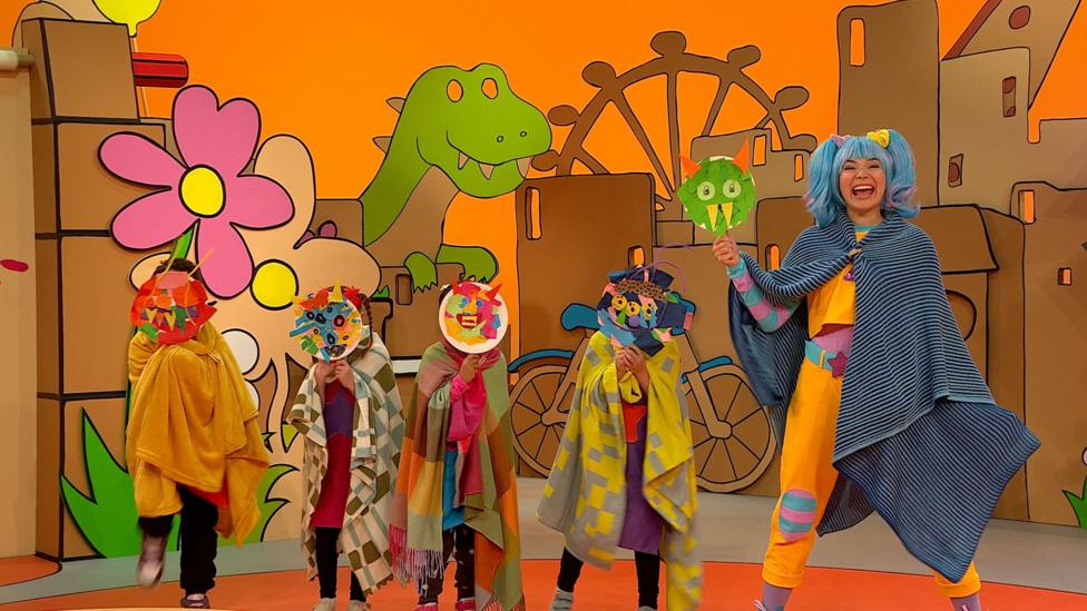 Sketch, a blue-haired woman, wears a cape and holds a monster mask made from a paper plate, as do some children next to her, pulling scary faces.