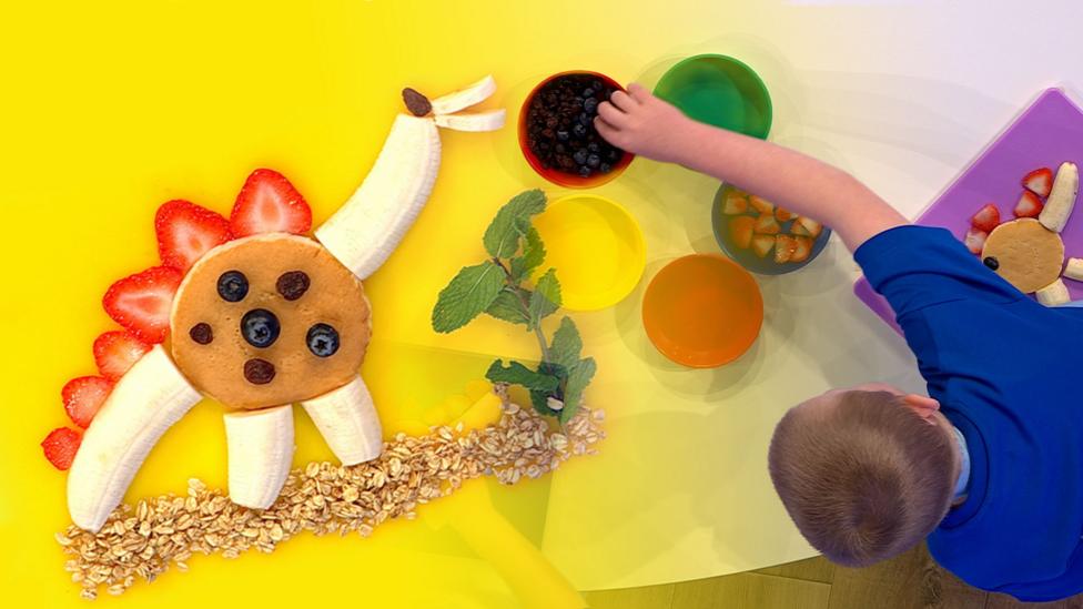 A boy leans over, constructing a dinosaur from a banana, a pancake and blueberries.