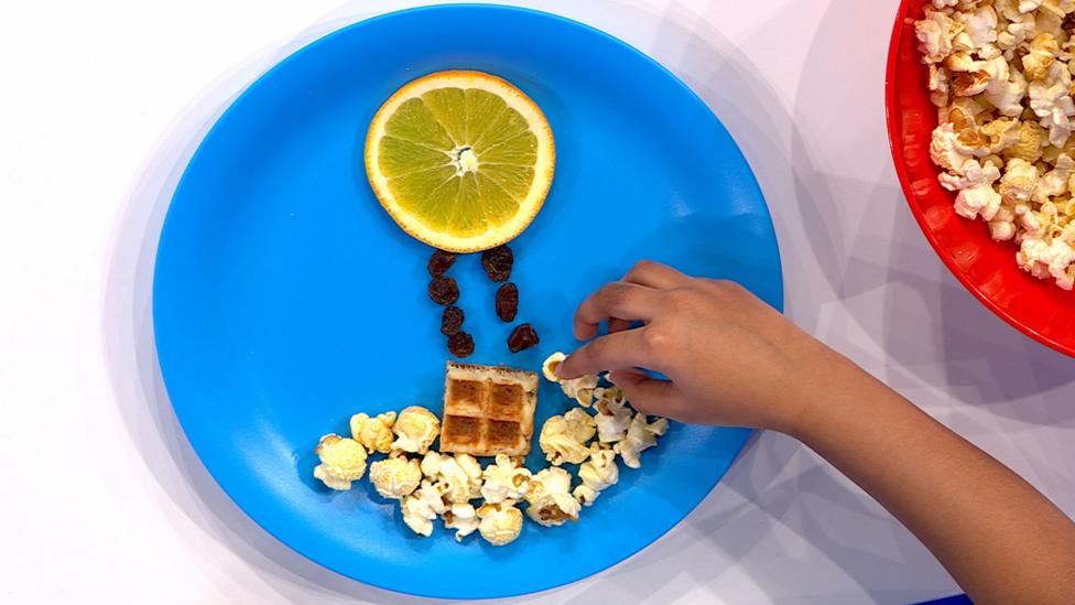 A blue plate featuring a hot air balloon shape made from a slice of orange, raisins and a piece of waffle, with popcorn for clouds.
