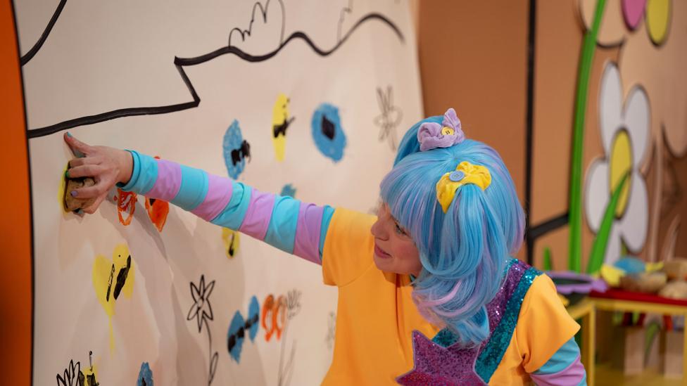 Sketch, a blue-haired woman, uses a potato print to stamp stripes on yellow circles on a wall mural, making bees.