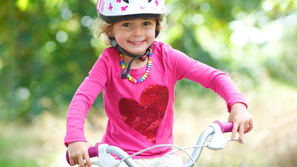 A little girl sat on a bike and wearing a bicycle helmet. She is smiling.