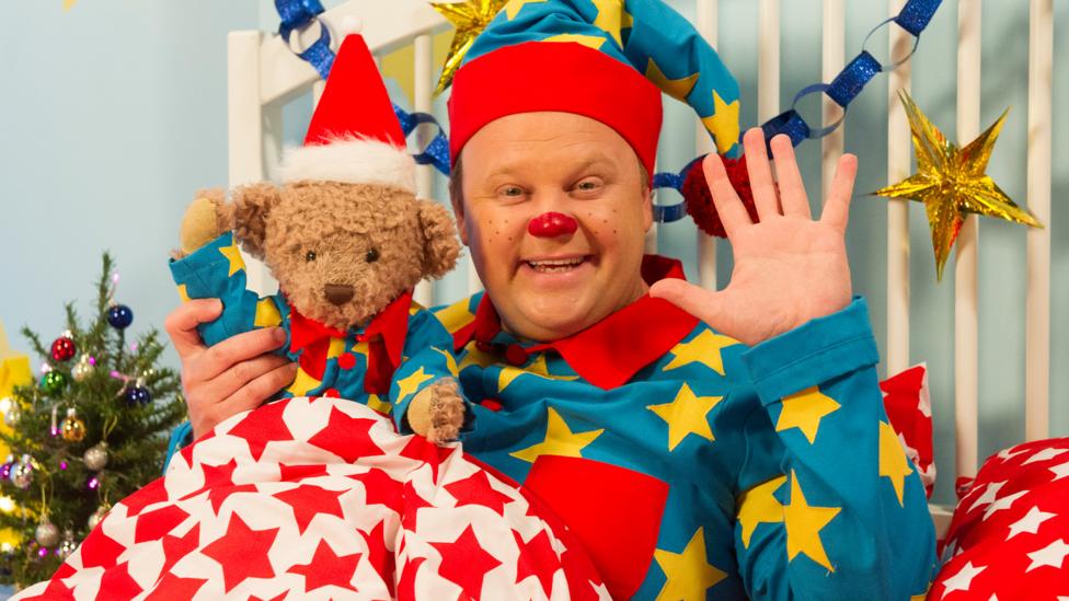 Image of a man waving dressed as a Christmas elf and holding a teddy bear in a red Christmas hat