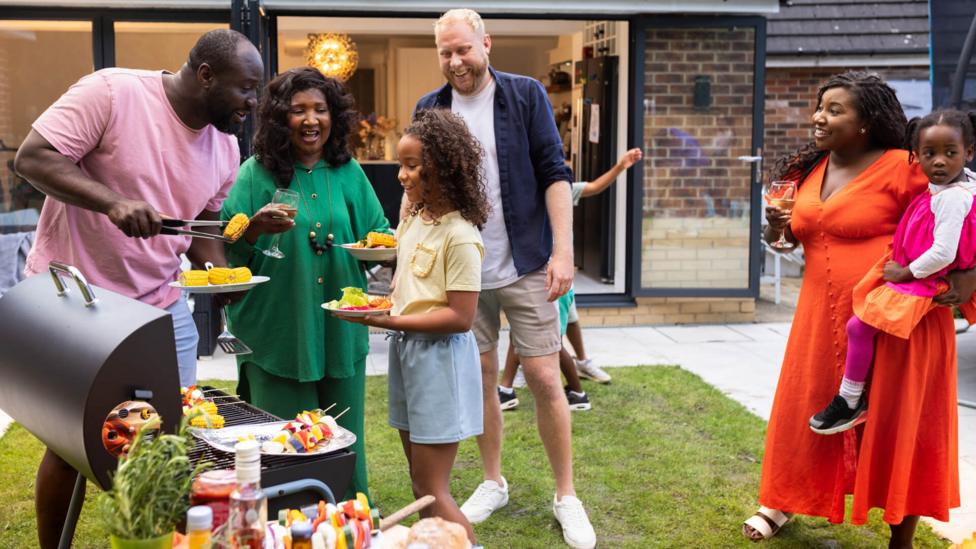 Black family featuring three generations plus family friends in back yard with BBQ, smiling and chatting.
