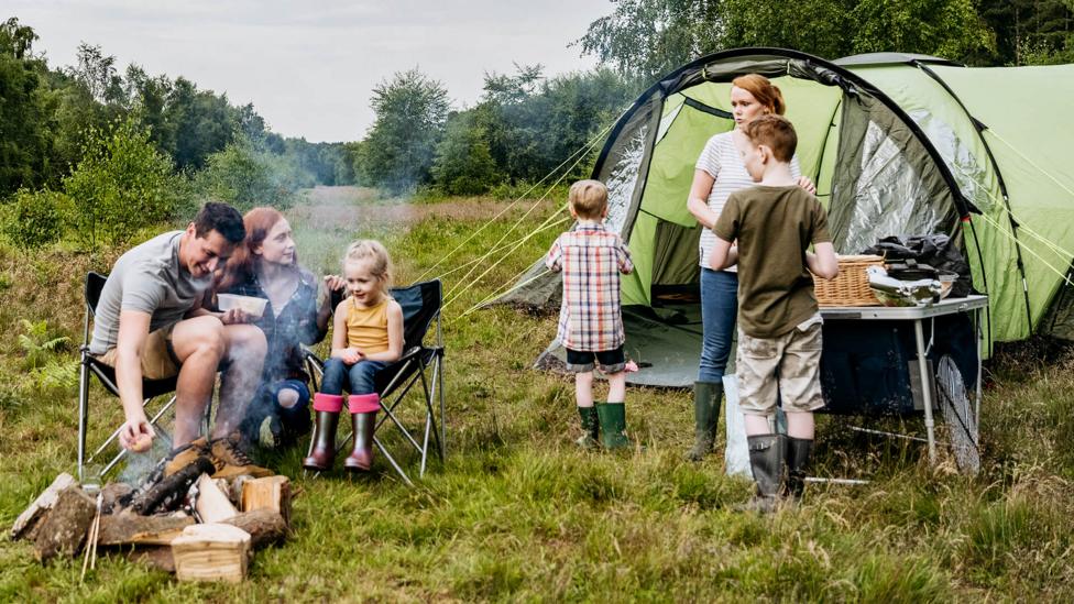 Family on a campsite, children gather around the tent and campfire with parents toasting marshmallows.