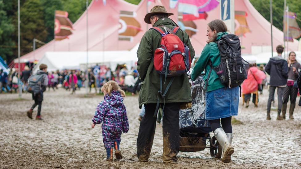 A toddler wearing a rain suit walking next to her father and mother. The mother is pushing a pram as they walk through a very muddy field, with big tents in the background.