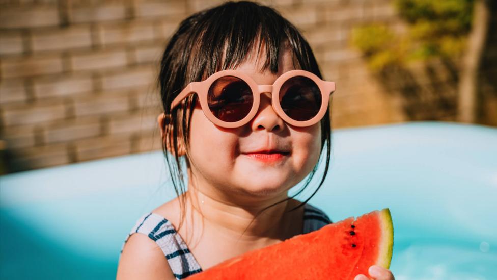 Asian little girl wearing sunglasses, sat in a paddling pool enjoying watermelon slices.