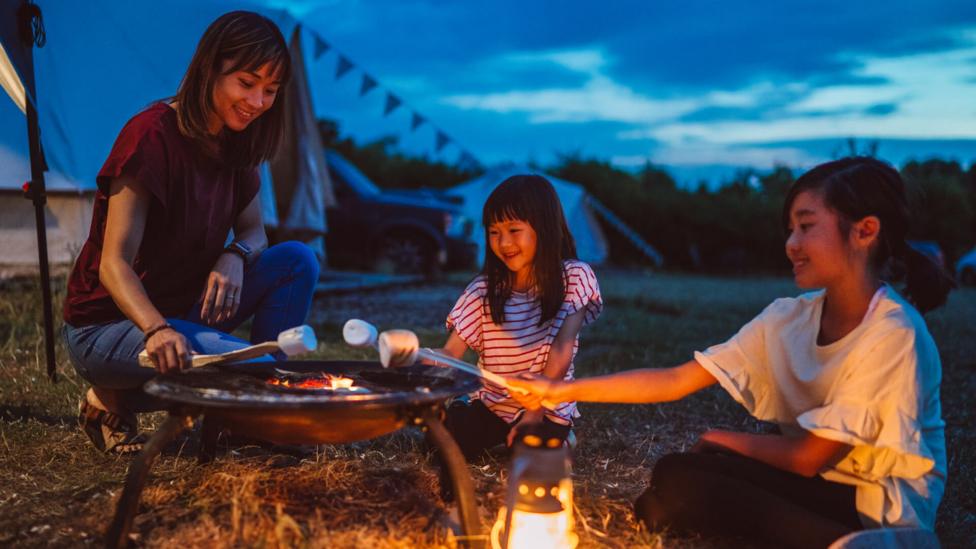 Asian mum with two little girls toasting marshmallows at campgrounds by a firepit, next to tents as the stars come out.