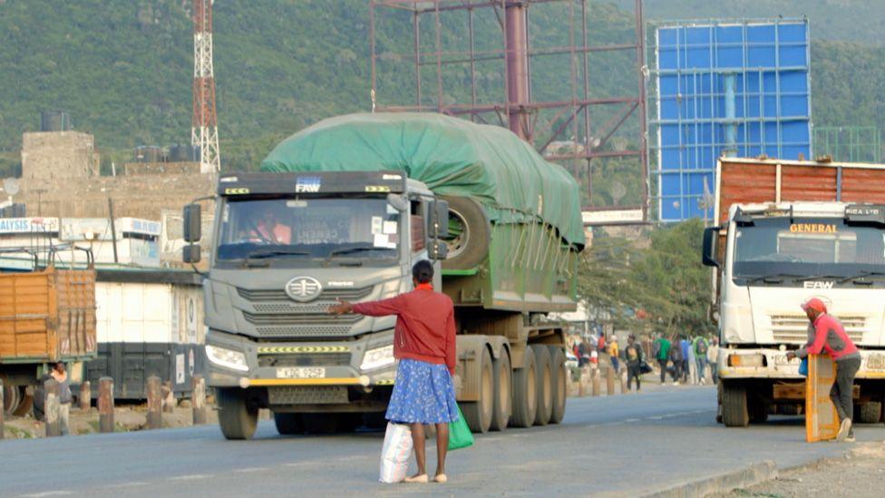 A street pictured during the day in Maai Mahiu. Two lorries can be seen on the tarmac road, another is parked to the side. One woman, in a blue dress and red jacket, puts out her hand to hitch a lift. Other pedestrians can be seen in the background.