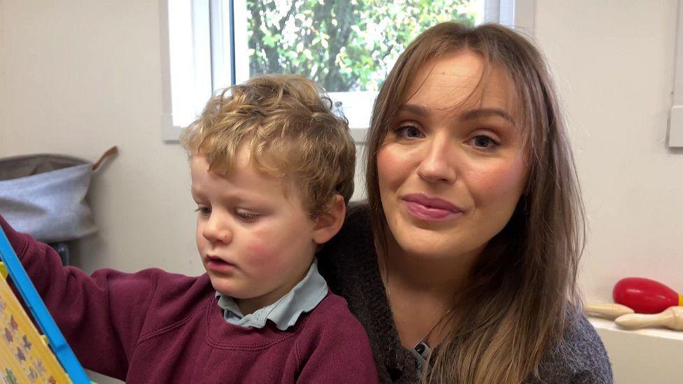 Sophie and her son Reggie sat in a waiting room of a doctor's surgery. Sophie has long brown hair and her son has blonde hair, is wearing a purple jumper and is playing with a toy.