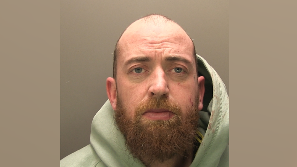 A police mugshot of Samuel Simpkins. He is bald with a light brown beard. He is wearing a green hoodie.