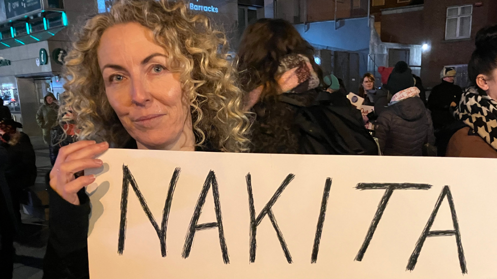 A woman with shoulder-length, blond, curly hair appears in close-up. She is holding a sign although the words cannot be seen in the image. She is standing outside with other people during a protest.