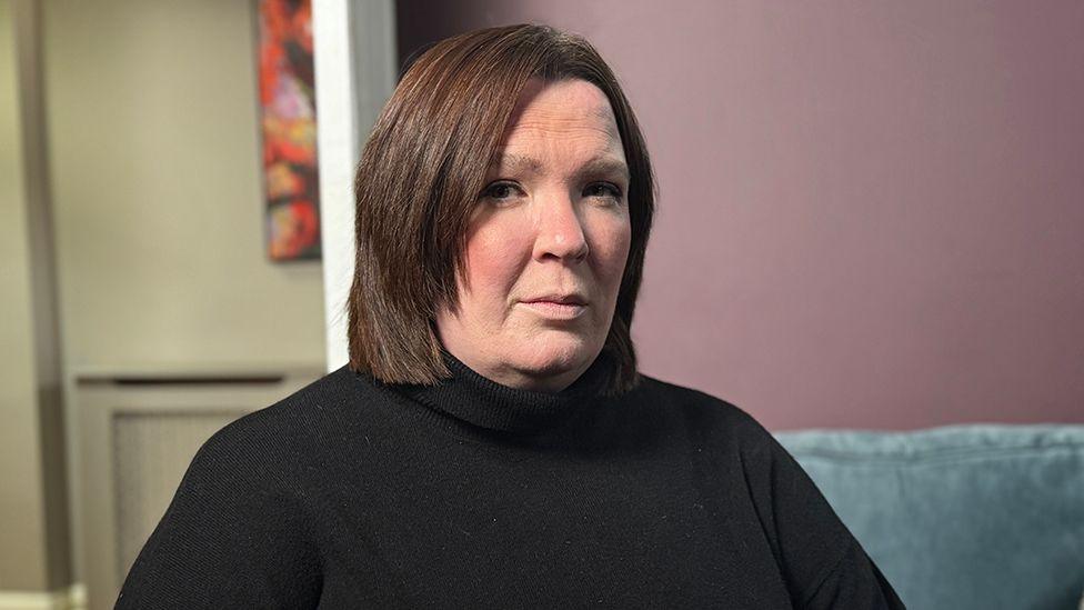 Catriona is wearing a black turtleneck sweater is seated indoors on a light blue couch. The background features a mauve-colored wall with a colorful abstract painting partially visible on the left side, and a doorway leading to another room. The lighting is soft and even, creating a calm atmosphere.