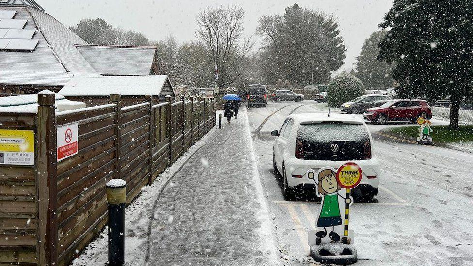 Wooden fence, path and road with school road signs, several parked cars and snow falling 