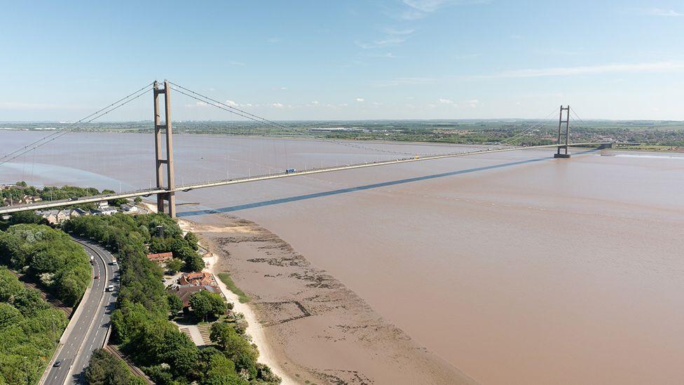 Aerial view of the Humber Bridge from the north bank of the Humber Estuary showing the bridge spanning the muddy water with vehicles crossing and traffic on the A63 road by the side of the bridge