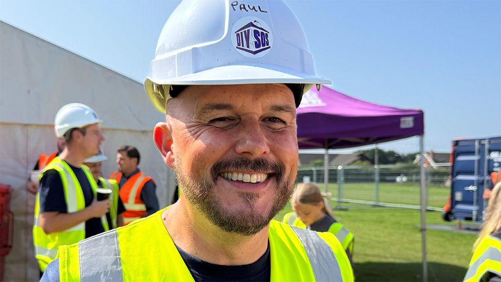 Close up of Paul Taylor in the sunshine smiling in a white hard hat and yellow high vis vest. He has a short dark beard
