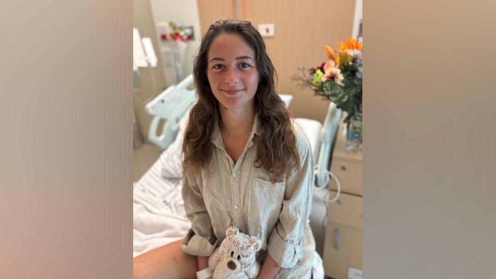 Caroline Wilga sits in a hospital bed with a teddy on her lap. She has long brown hair and is smiling at the camera.