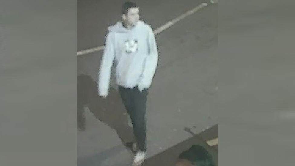 CCTV of man standing in a street wearing a pale hoodie and dark trousers