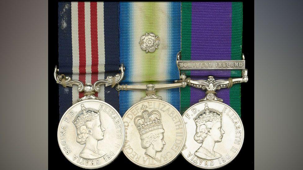 Three silver medals in a row and their ribbons. Left to right Military Medal, on a red white and blue ribbon; South Atlantic medal with a rosette on a blue cream and lime green ribbon; General Service medal for Northern Ireland on a green and purple ribbon. 