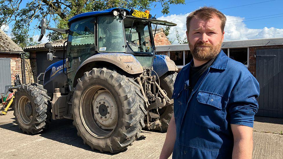 Farmer John Storey standing next to a tractor wearing blue overalls