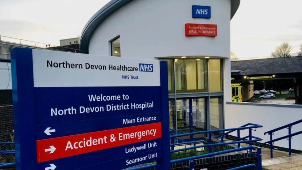 A sign which reads: Northern Devon Healthcare NHS Trust, Welcome to North Devon District Hospital, and there are arrows pointing to the Main Entrance, Accident and Emergency, Ladywell Unit and Seamoor Unit. There is a building behind the sign with a double door.