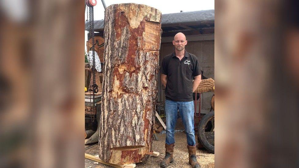 A man with no hair wearing a black t-shirt stood next to a 7ft 3in wooden log in Whitwell in Derbyshire