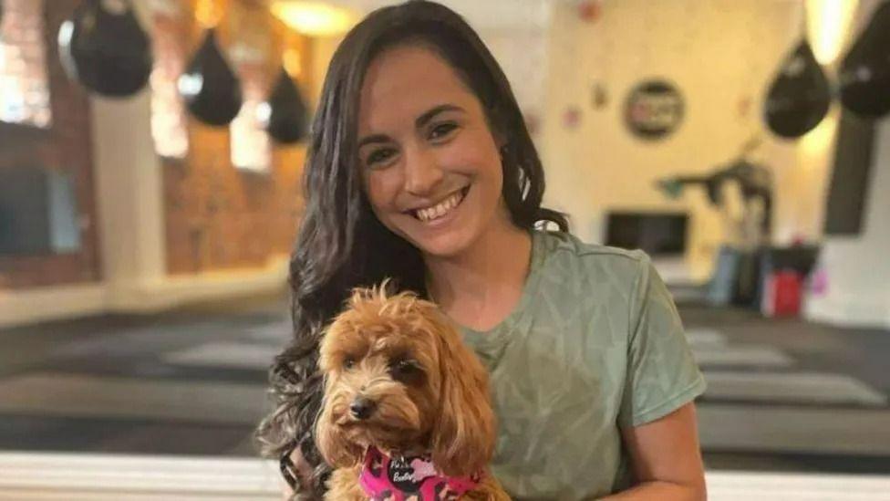 Leanne Lucas in a yoga studio surrounded by yoga mats. She's holding a small dog. She wears her long brown hair down in waves and is wearing a pale green t-shirt as she smiles at the camera. 