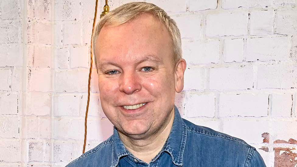 Head and shoulders image of Steve Pemberton smiling in front of a white painted brick wall and wearing a denim shirt