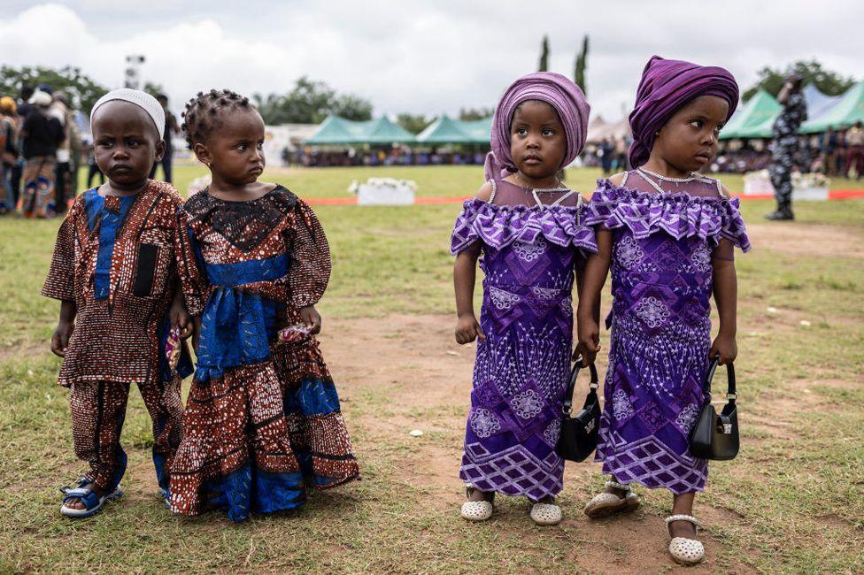 Two children wearing brown patterned clothing stand near two children wearing purple pattern clothing. Green tents can be seen in the distant background on October 12, 2024.