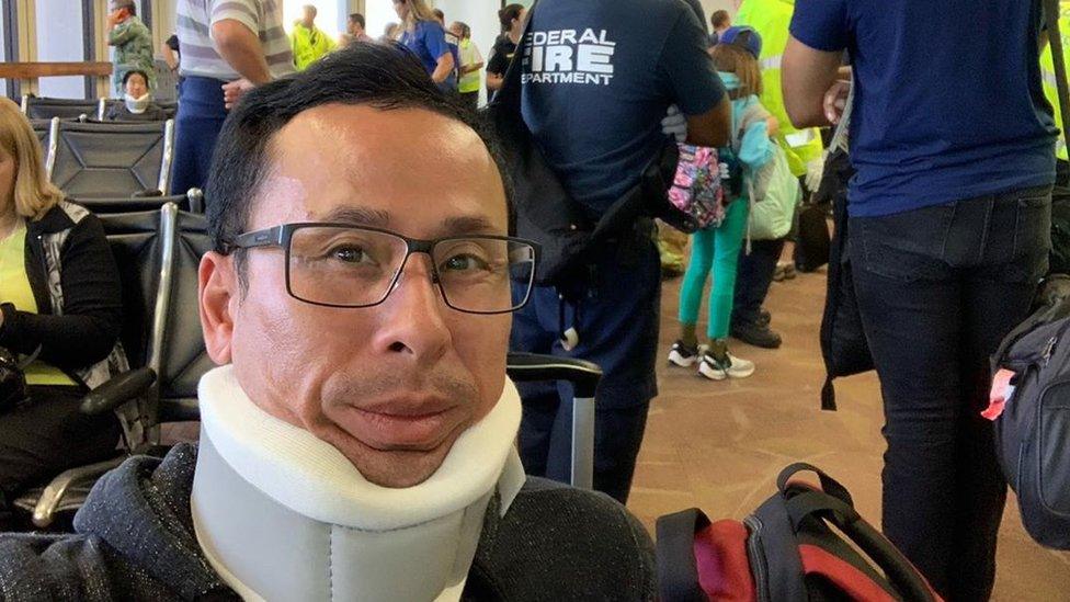 A man takes a selfie wearing a neck brace, with fire department and others behind