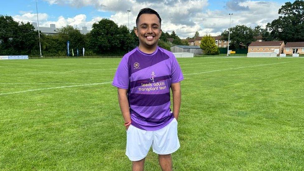 A young Asian man in a t-shirt, with an alternating light/dark purple pattern, stands in the middle of a sports field. His t-shirt says "Leeds adults transplant team" in gold letters. He's smiling with his hands in his white shorts pockets. The field around him looks immaculately well-kept, with white lines painted on it. In the distance a small, beige clubhouse building is visible.