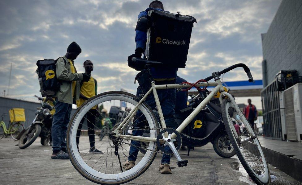 A group of men standing with motorcycles parked around. One man is sitting on a bicycle.