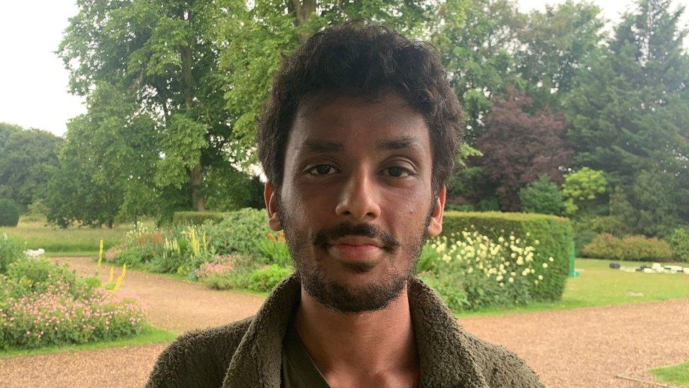 Bilal Ally stood in a garden. He has dark brunette hair and a beard and is wearing an olive green fleece. Behind him there are trees and a hedgerow and various flowerbeds separated by stone pathways.