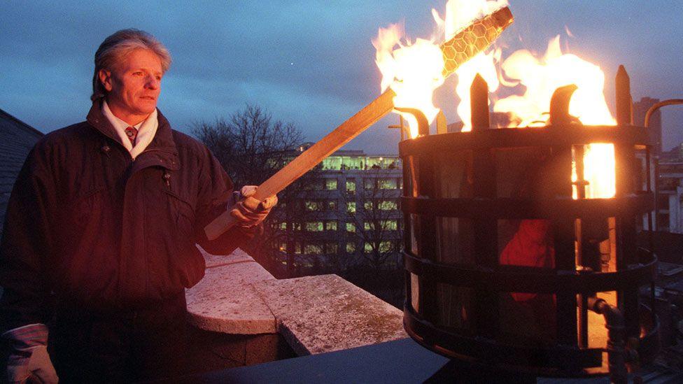 A grainy photograph of Bruno Peek lighting a beacon on the tower of St James's Church in Clerkenwell Green, London to mark the new Millennium. He is standing on the left and his holding a beacon in his left hand. It is lighting a metal beacon stand which is on the right. Behind it can be be seen an mid-century office or flat building