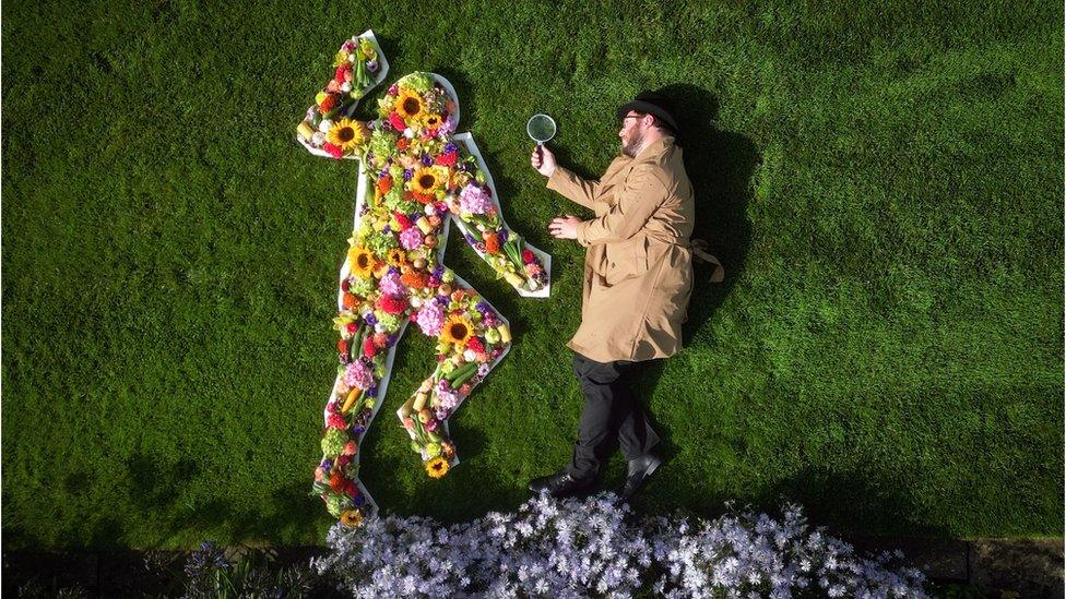 Alexander McDonnell, playing the role of a detective, views a floral crime scene figure that forms part of a floral art installation